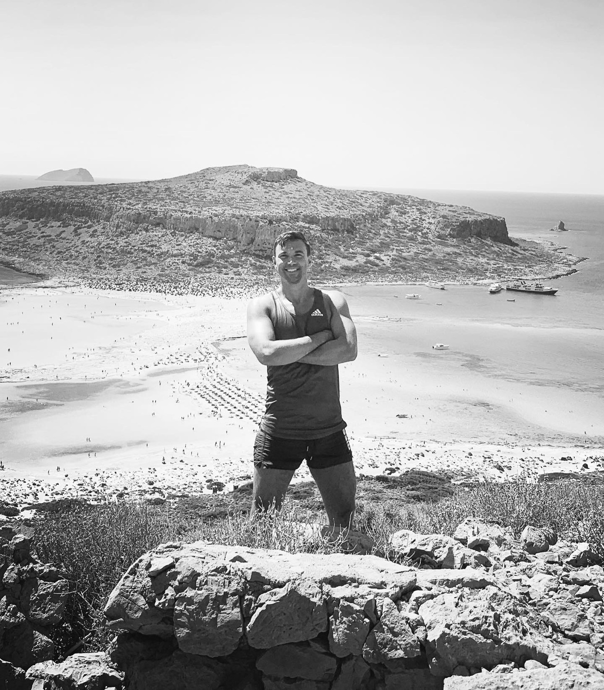 A muscular man poses with his arms crossed on a rocky outcrop overlooking a crowded beach and the ocean. The black and white photo captures a scenic coastal landscape with distant hills and boats.