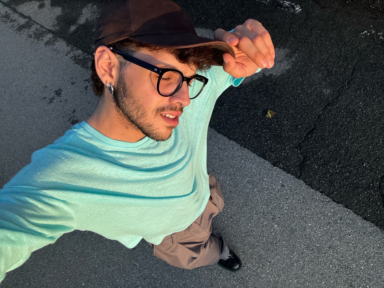 A man takes a selfie, looking down at the camera. He wears glasses, a cap, and a light blue long-sleeve shirt.