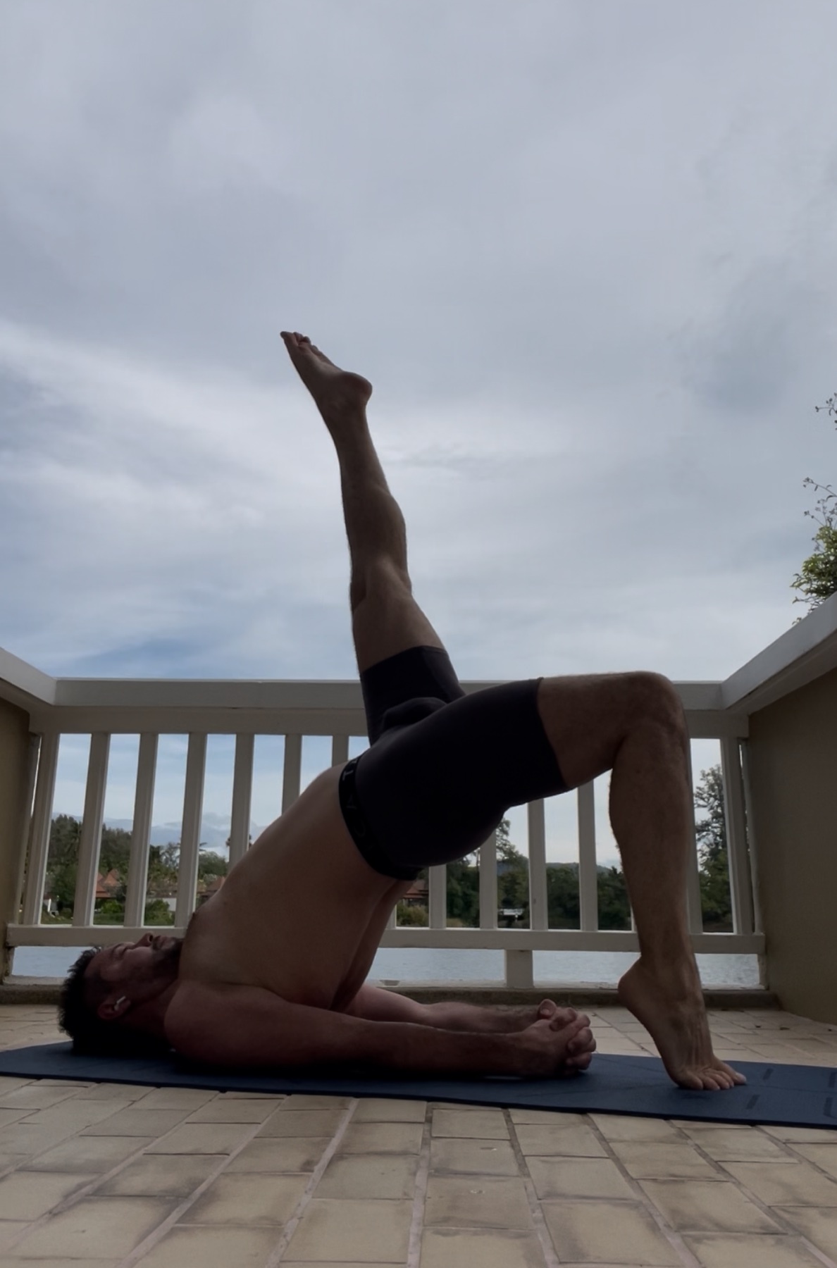 A man is doing yoga on a mat outside. He is shirtless and flexing his muscular legs.