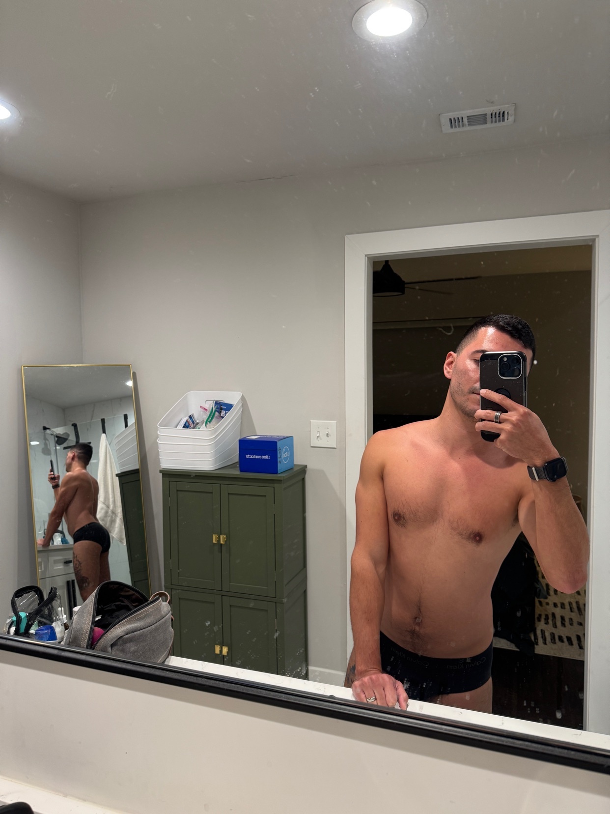 A man takes a mirror selfie, shirtless and in underwear. He flexes his arm and shows off his physique.