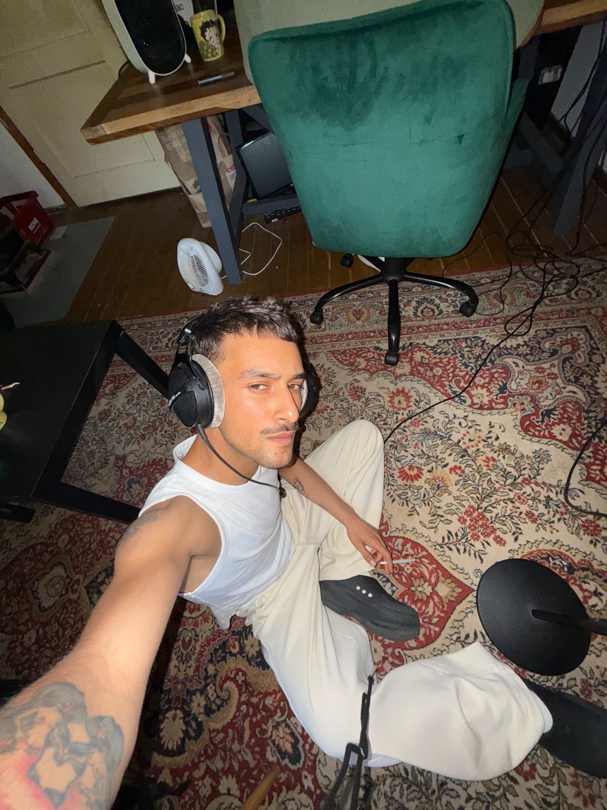 A man is taking a selfie while sitting on the floor in a studio. He is wearing a white tank top and headphones.
