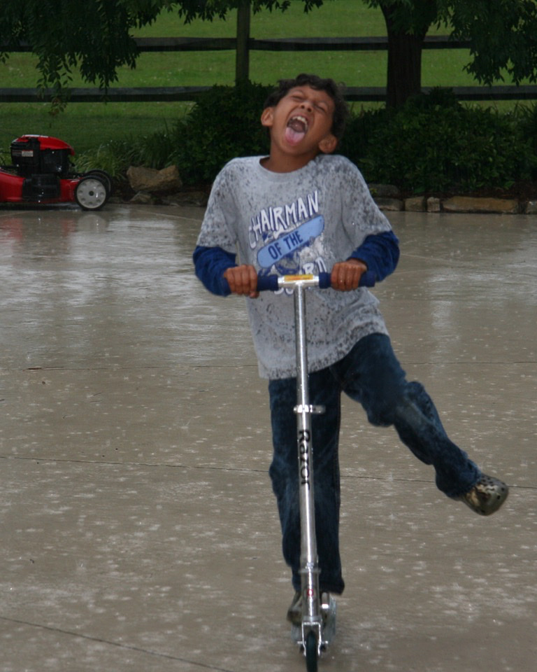 A young boy rides a scooter in the rain, with a joyful expression. He is wearing a t-shirt and jeans.