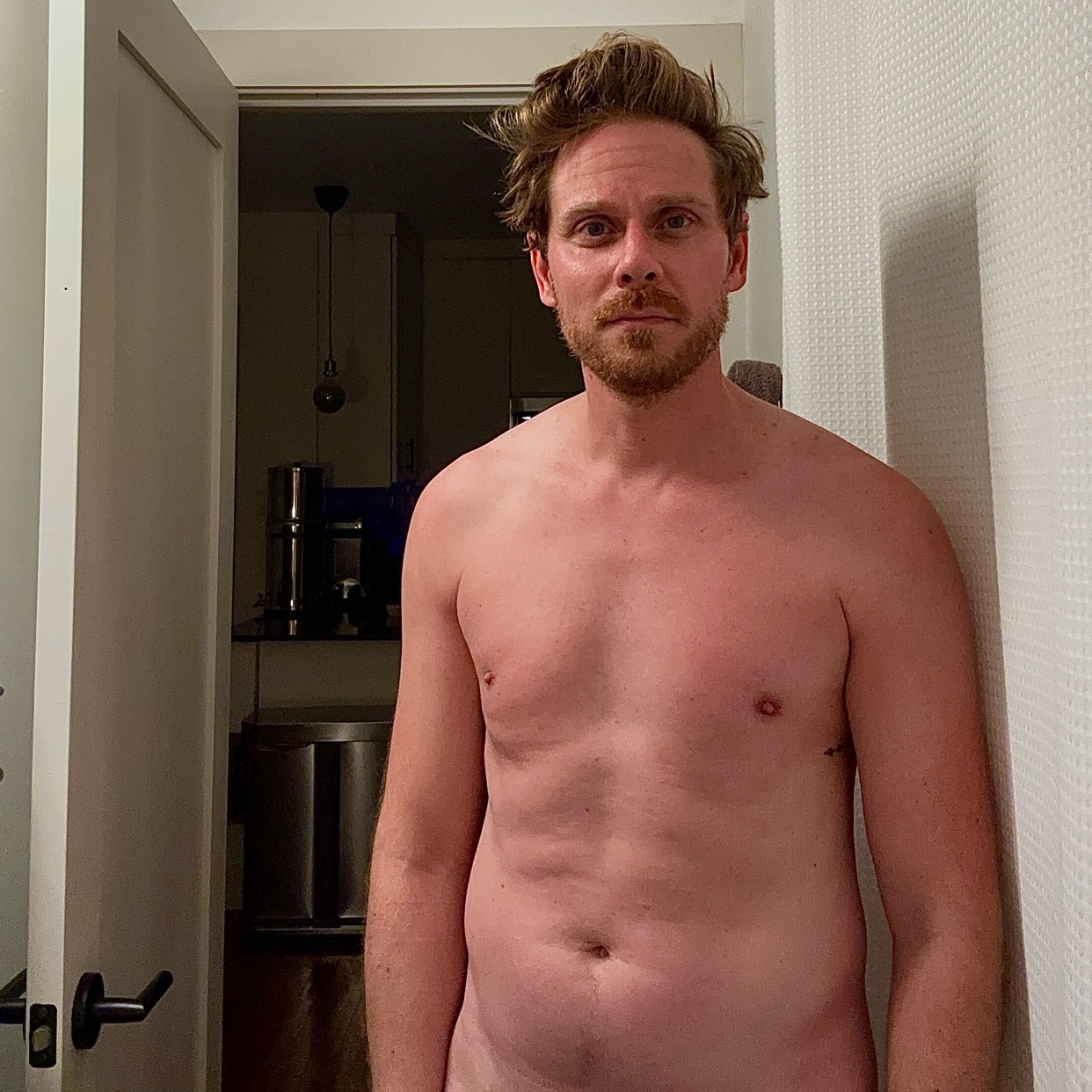 A man stands shirtless in a room, leaning against a wall. The photo is a selfie, with a door and kitchen visible in the background.