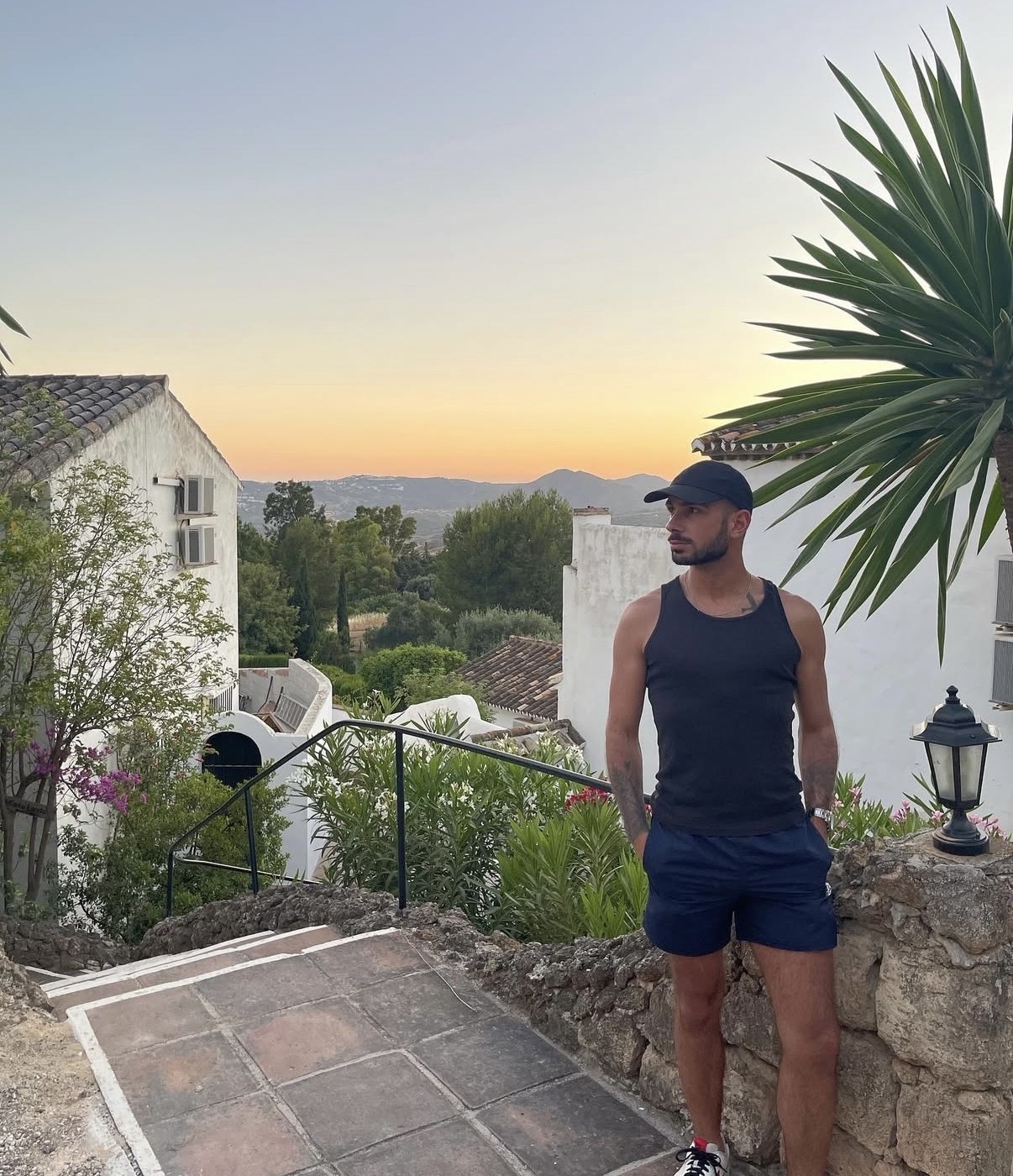 A man stands on a stone staircase overlooking a scenic landscape at sunset. He is wearing a tank top, shorts, and a baseball cap.