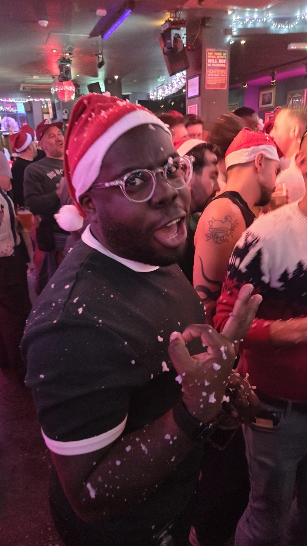 A man wearing a Santa hat and glasses is smiling and making an ok sign. The photo appears to be taken at a Christmas party.