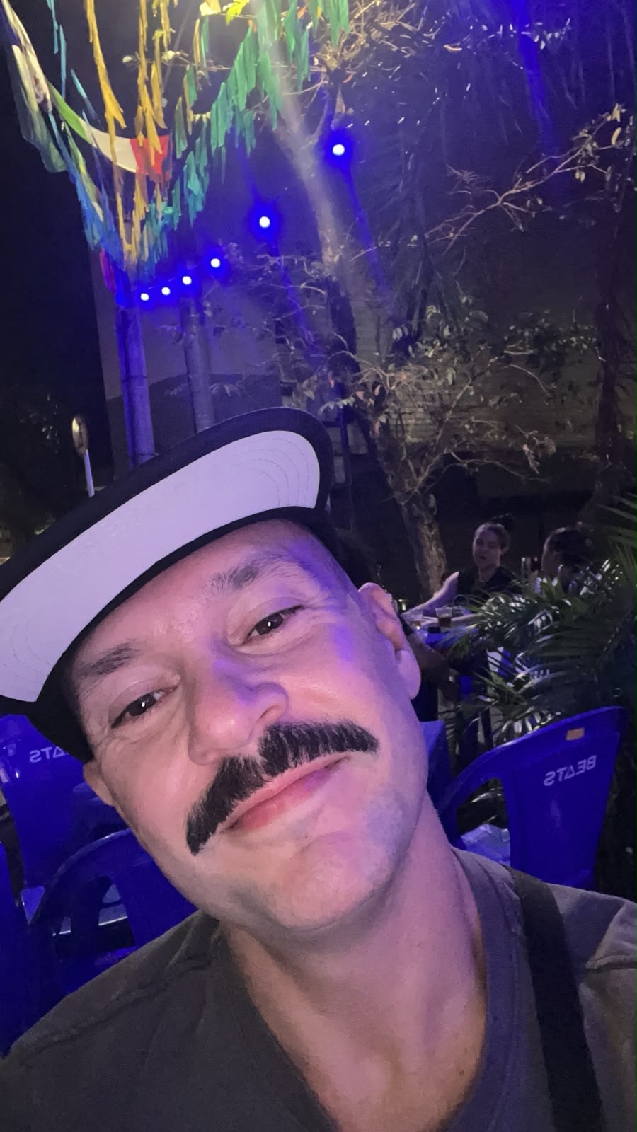 A man with a prominent mustache takes a selfie at night, with colorful party lights strung overhead and blurred figures in the background.