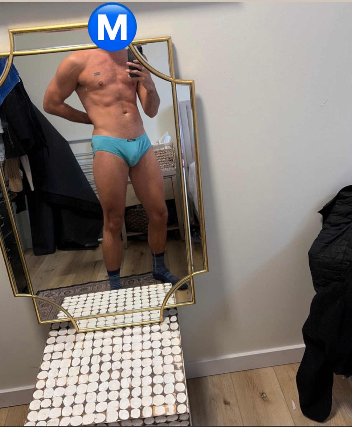 A man poses shirtless in front of a mirror, wearing blue underwear. The photo highlights his muscular physique.