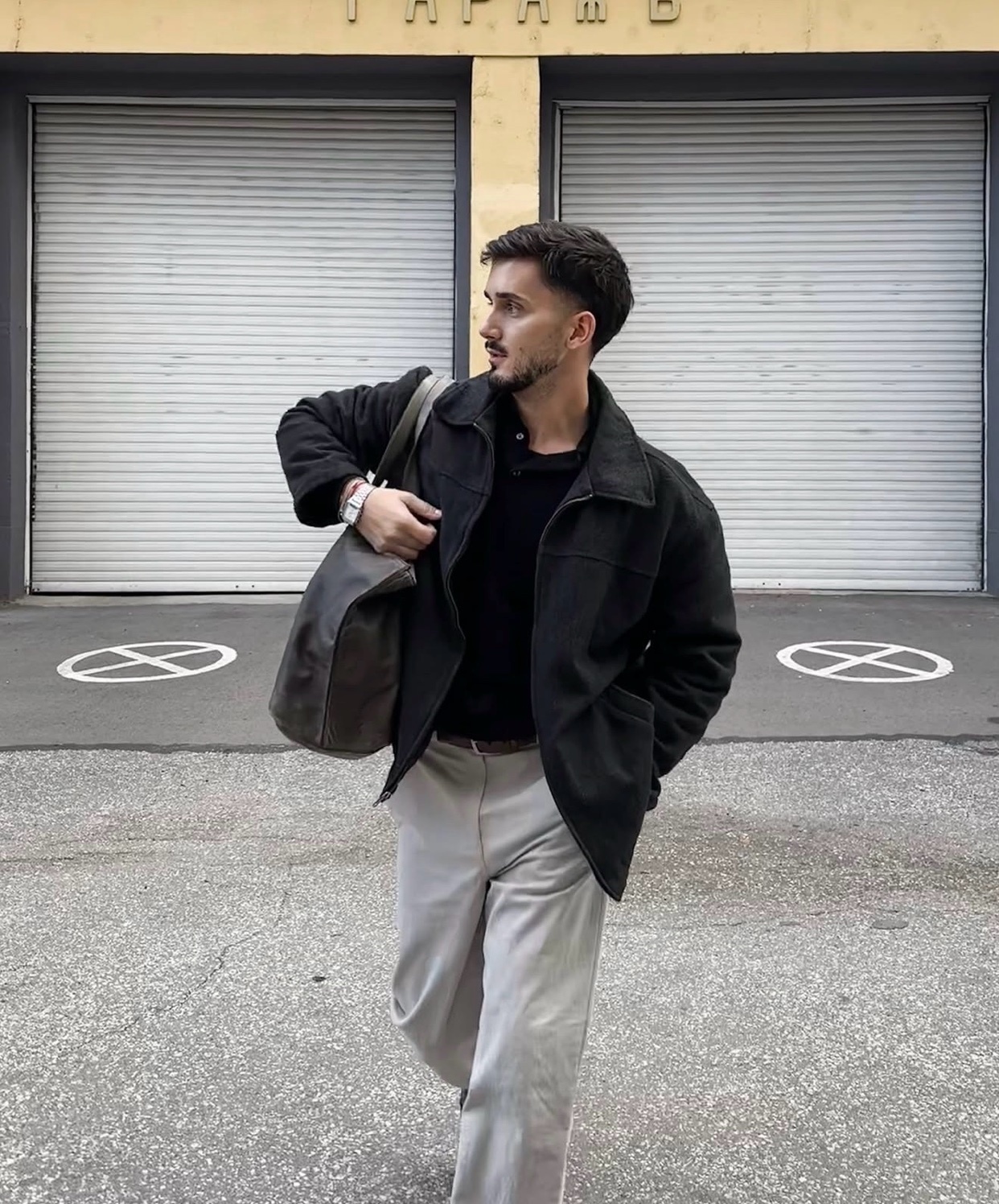 A man in a black jacket and light-colored pants walks with a large brown bag slung over his shoulder. He has dark hair and stubble, and is wearing a watch.