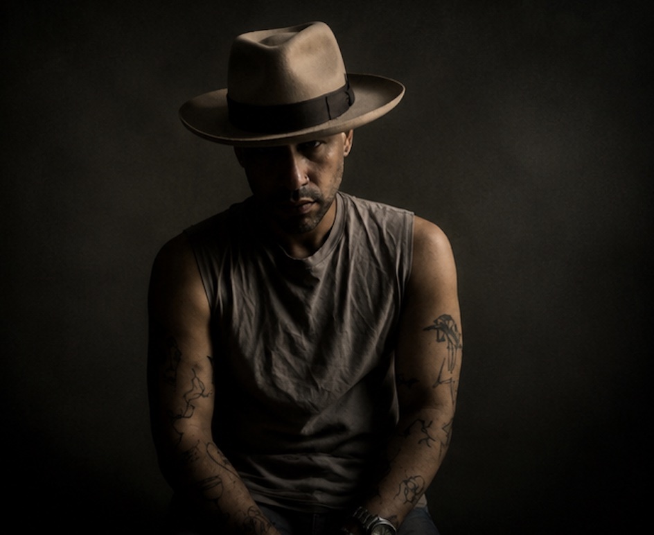 A man wearing a fedora and a tank top is pictured in a studio portrait. He has tattoos on his arms and is looking directly at the camera.