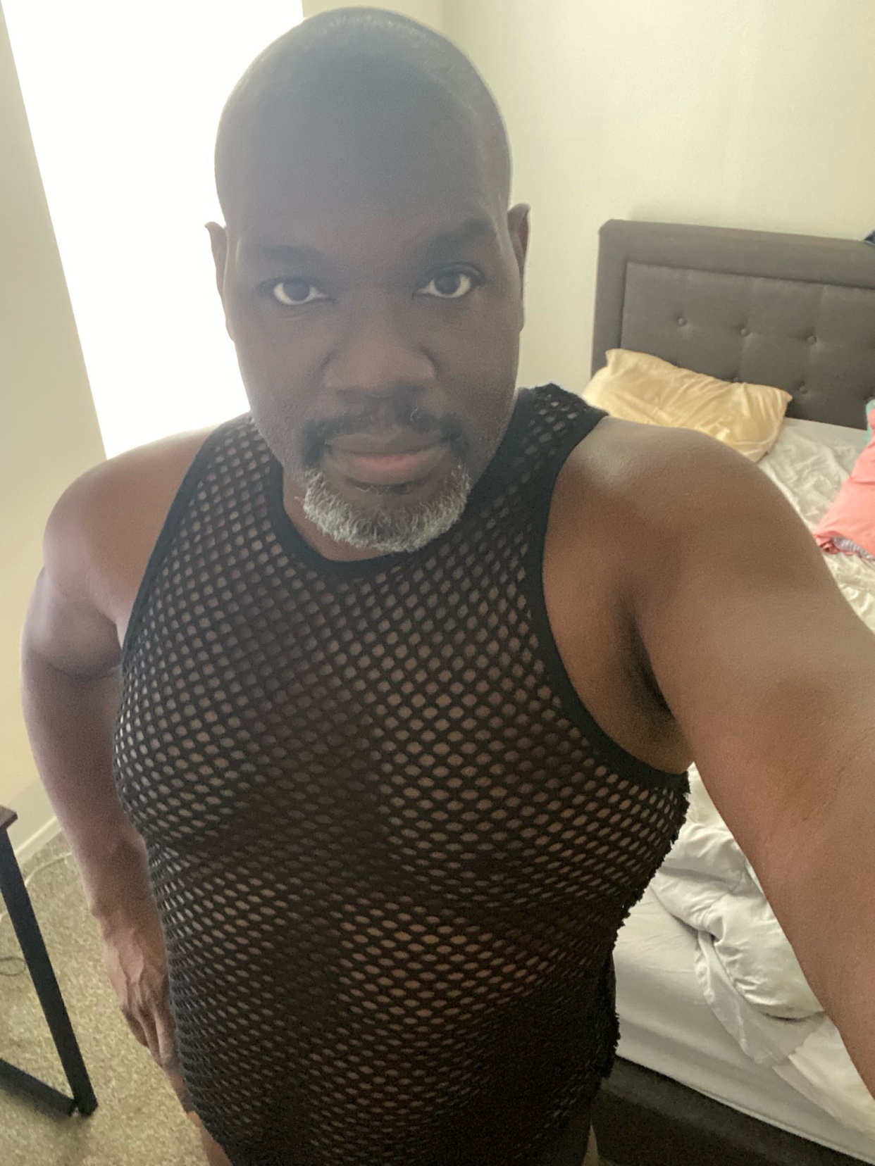 A man poses for a selfie in a mesh top. The photo is taken in a bedroom.