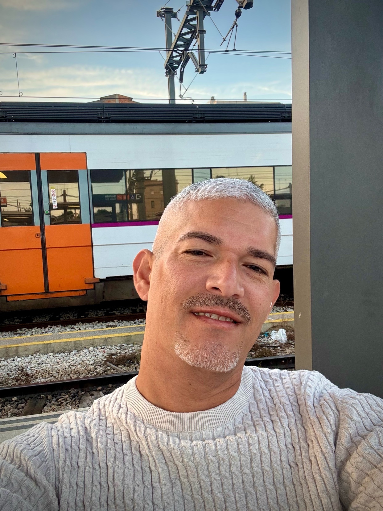 A man smiles for a selfie in front of a train. The photo is taken outdoors during the day.