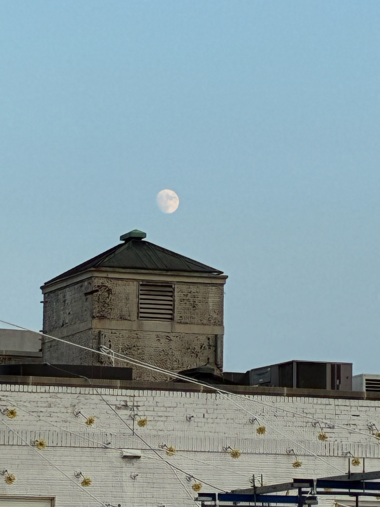 A full moon hangs in the clear sky above a weathered building with a distinctive square structure on its roof.