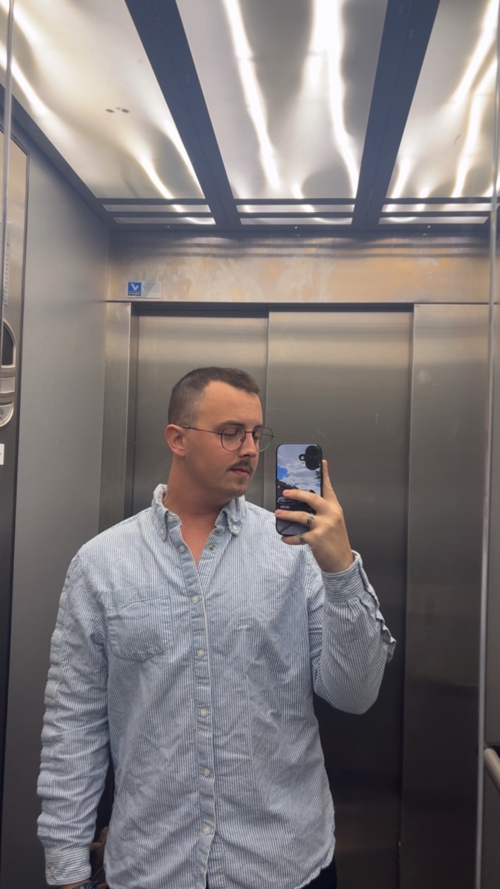 A man takes a selfie in an elevator, wearing glasses and a light blue button-up shirt.