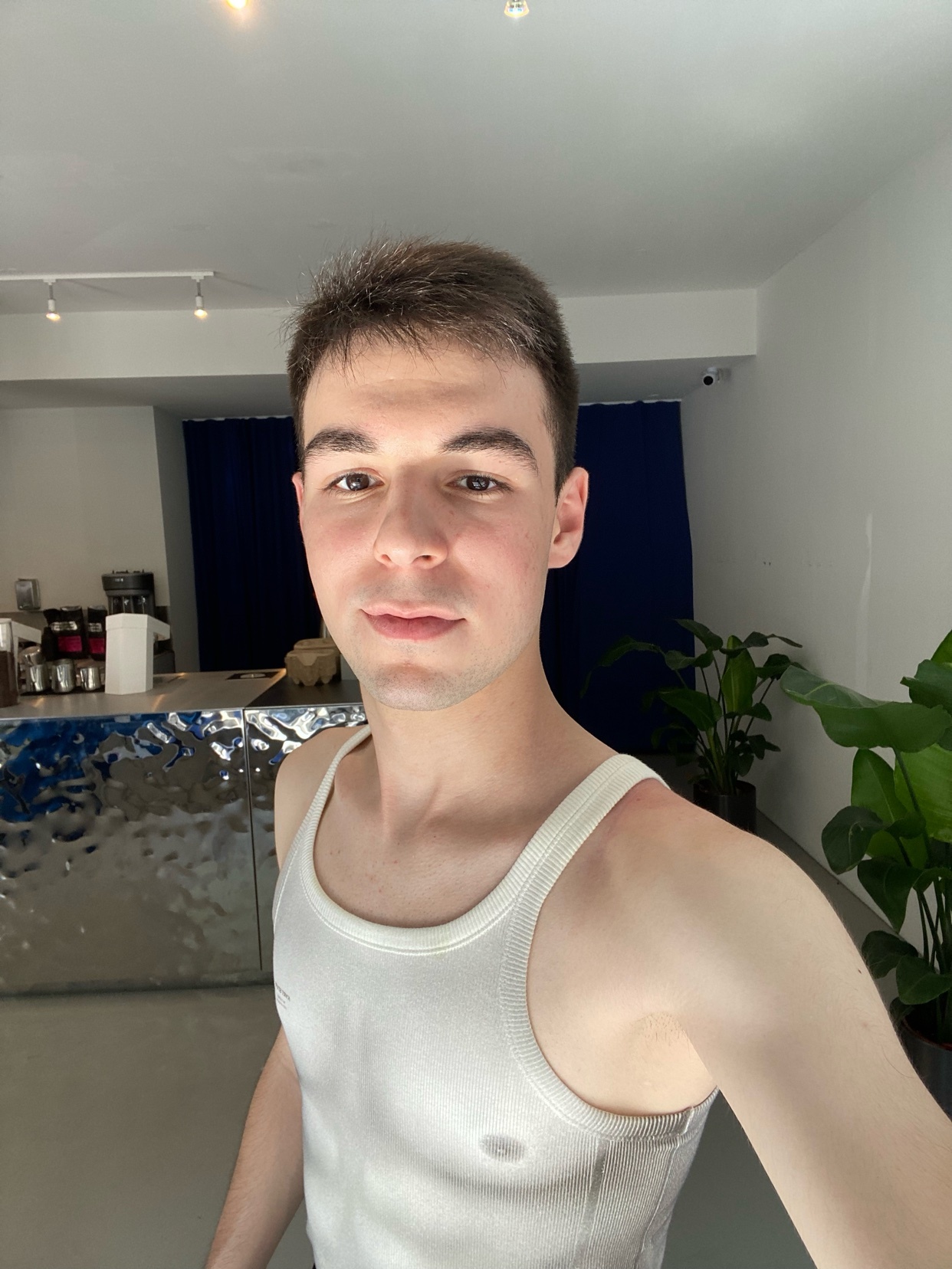 A man takes a selfie in a modern cafe. He is shirtless, wearing a white tank top.