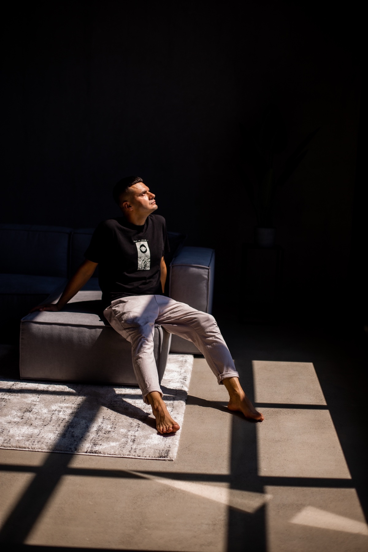 A man sits on a couch in a dimly lit room, bathed in dramatic sunlight and shadows. He is looking upwards, dressed in casual attire.