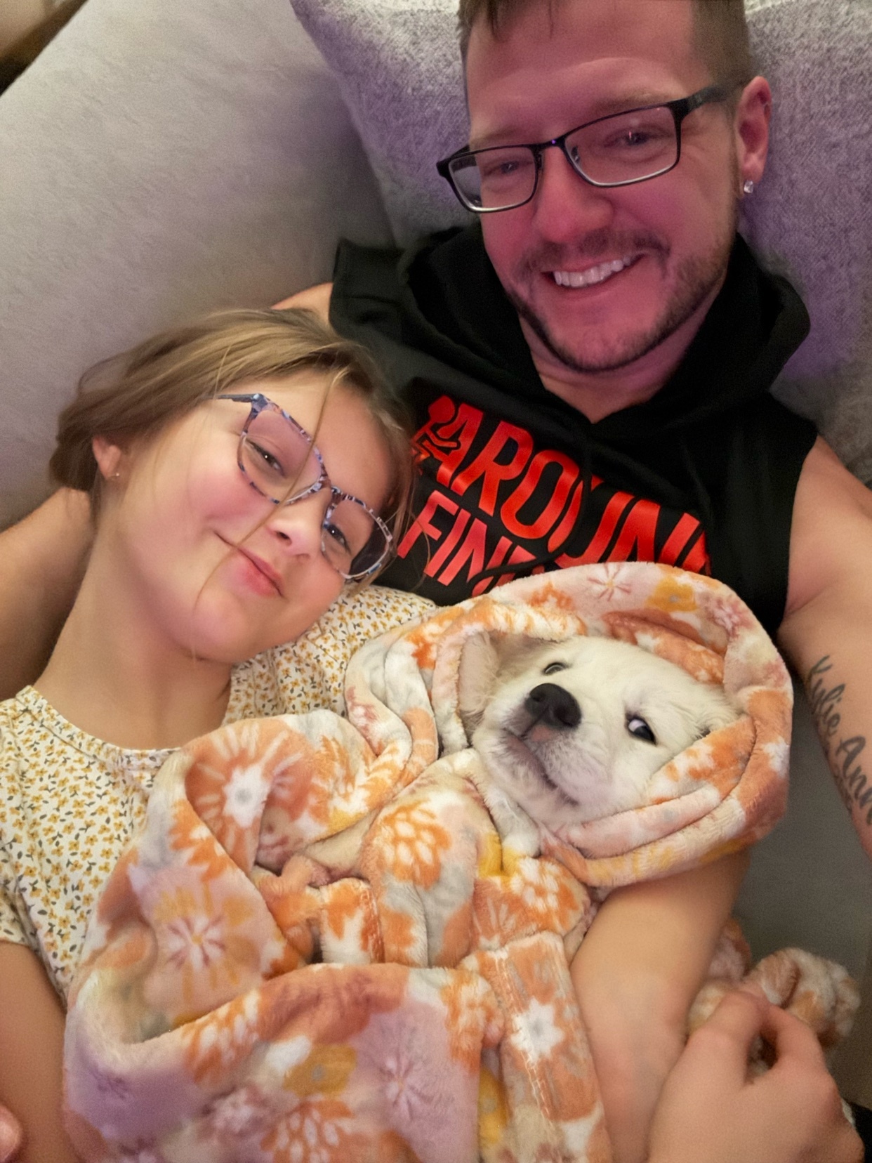 A man and a young girl smile as they cuddle a small white puppy wrapped in a floral blanket.
