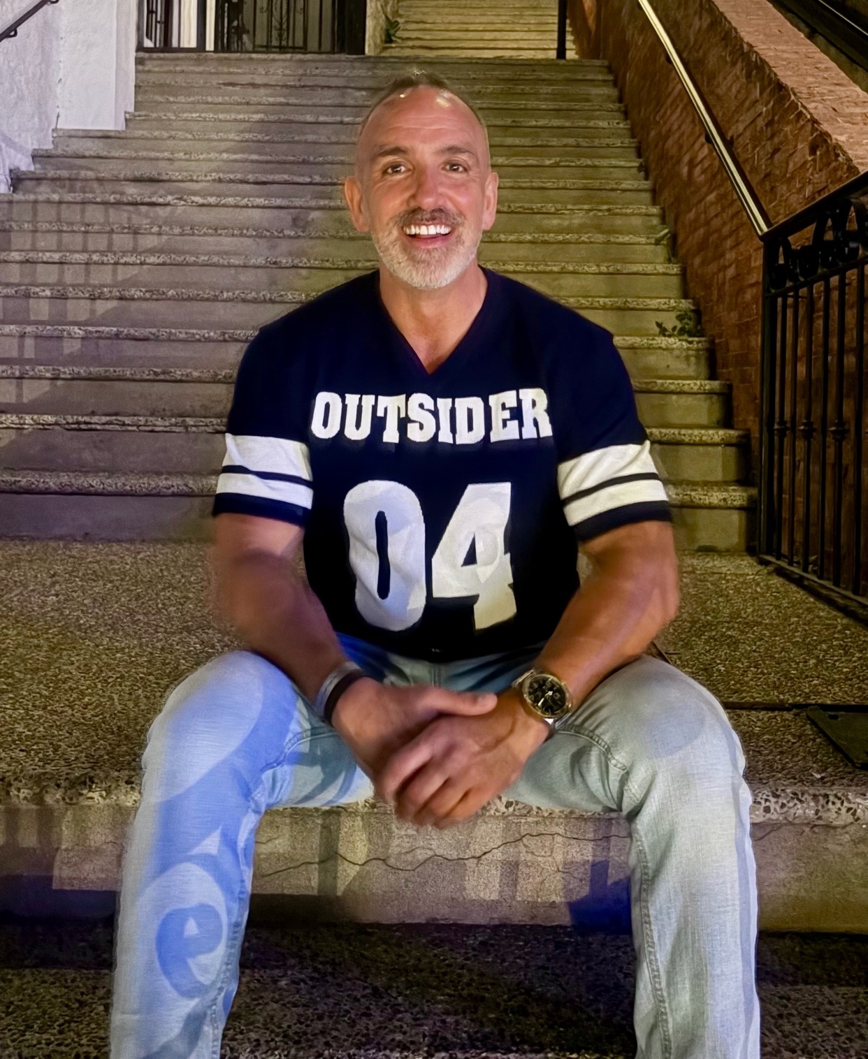 A man with a bald head and a beard smiles while sitting on concrete steps. He is wearing a navy blue t-shirt with white lettering that says "OUTSIDER 04" and light-wash jeans.