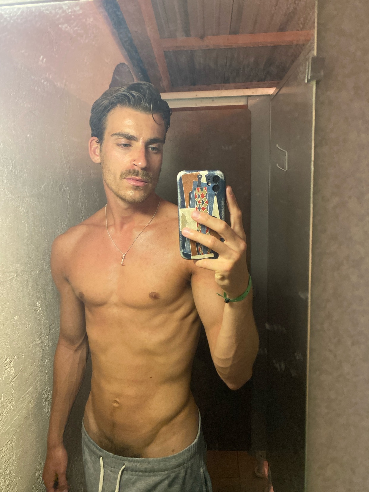 A shirtless man takes a mirror selfie. He has dark hair, a mustache, and is wearing gray sweatpants.