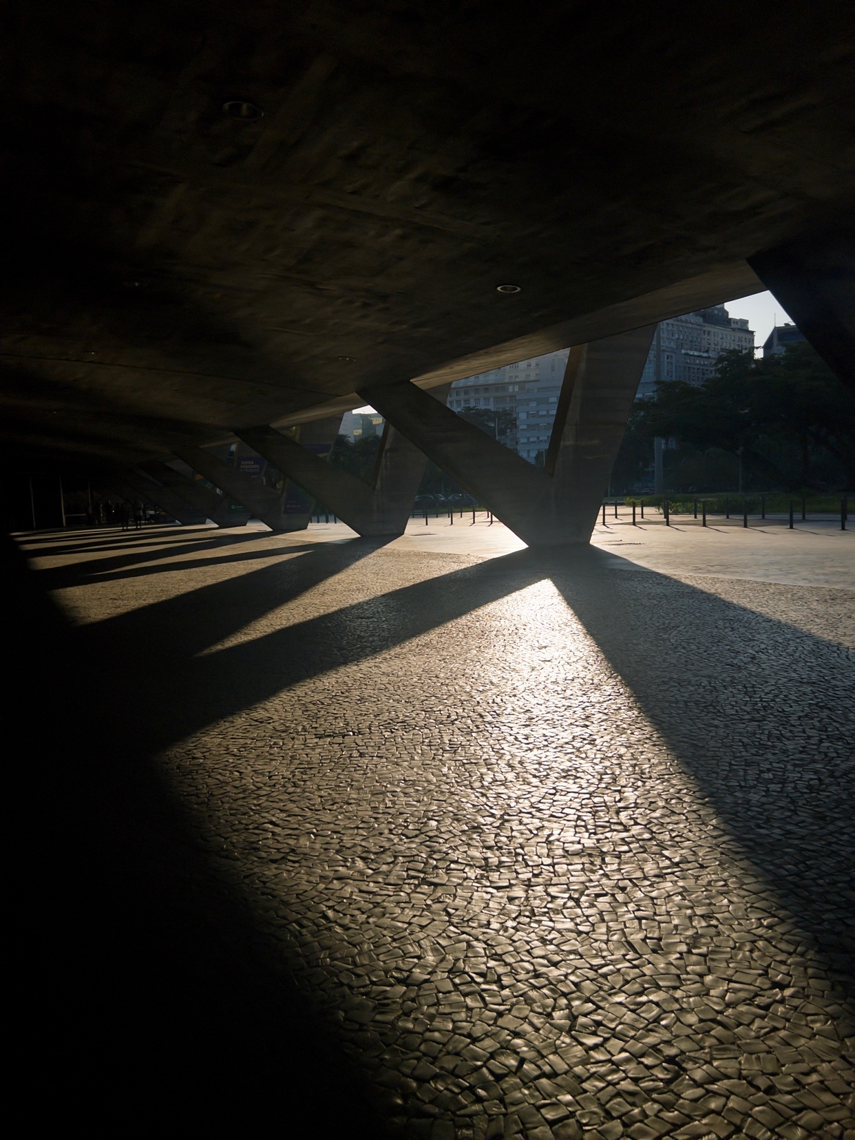 Sunlight streams through the geometric concrete supports of a modern building, casting dramatic shadows across a cobblestone ground. The interplay of light and shadow creates a striking visual pattern on the textured surface.