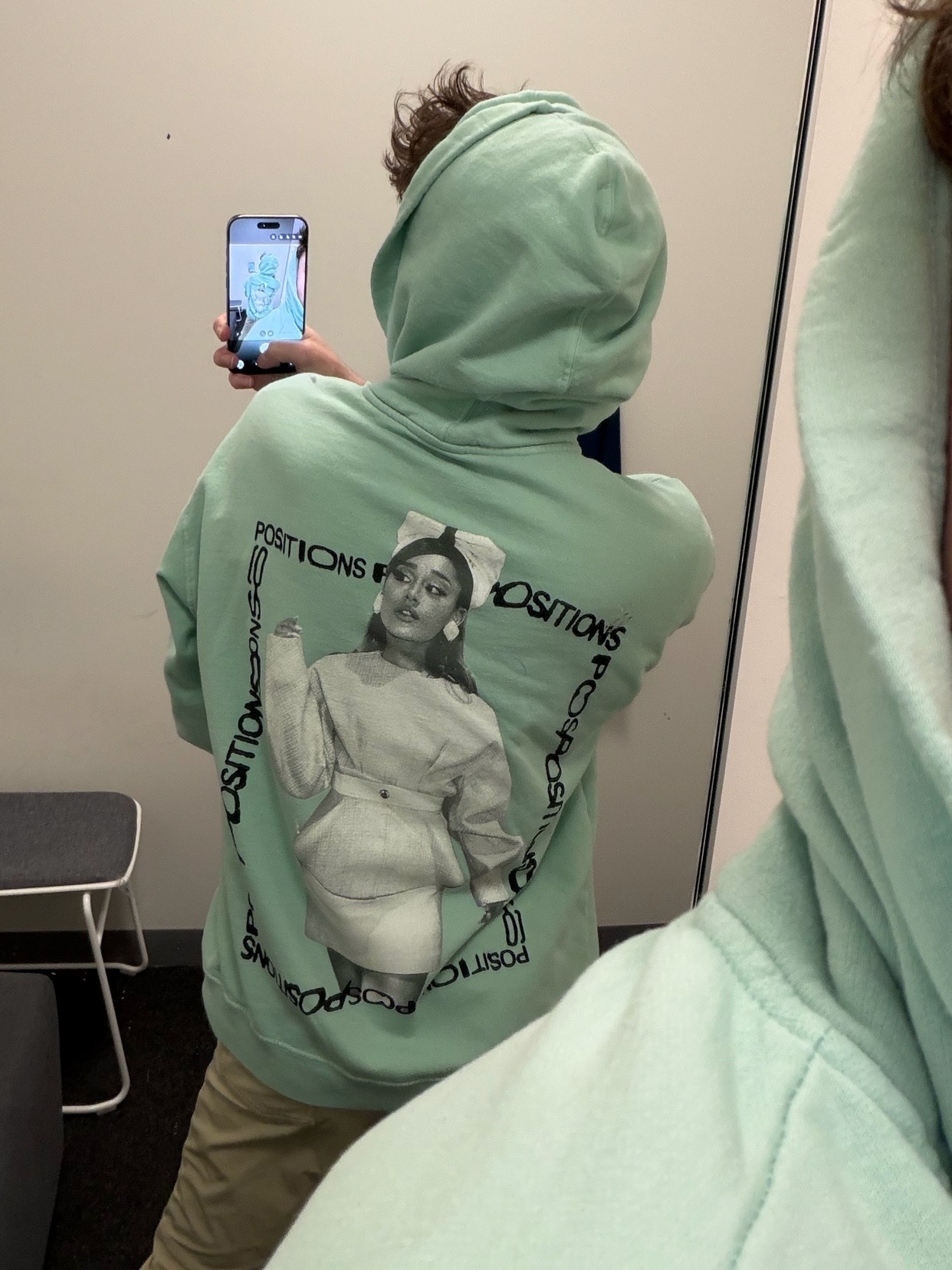 A person takes a mirror selfie wearing a green hoodie with Ariana Grande's "Positions" album art on the back. The photo is taken in a dressing room.