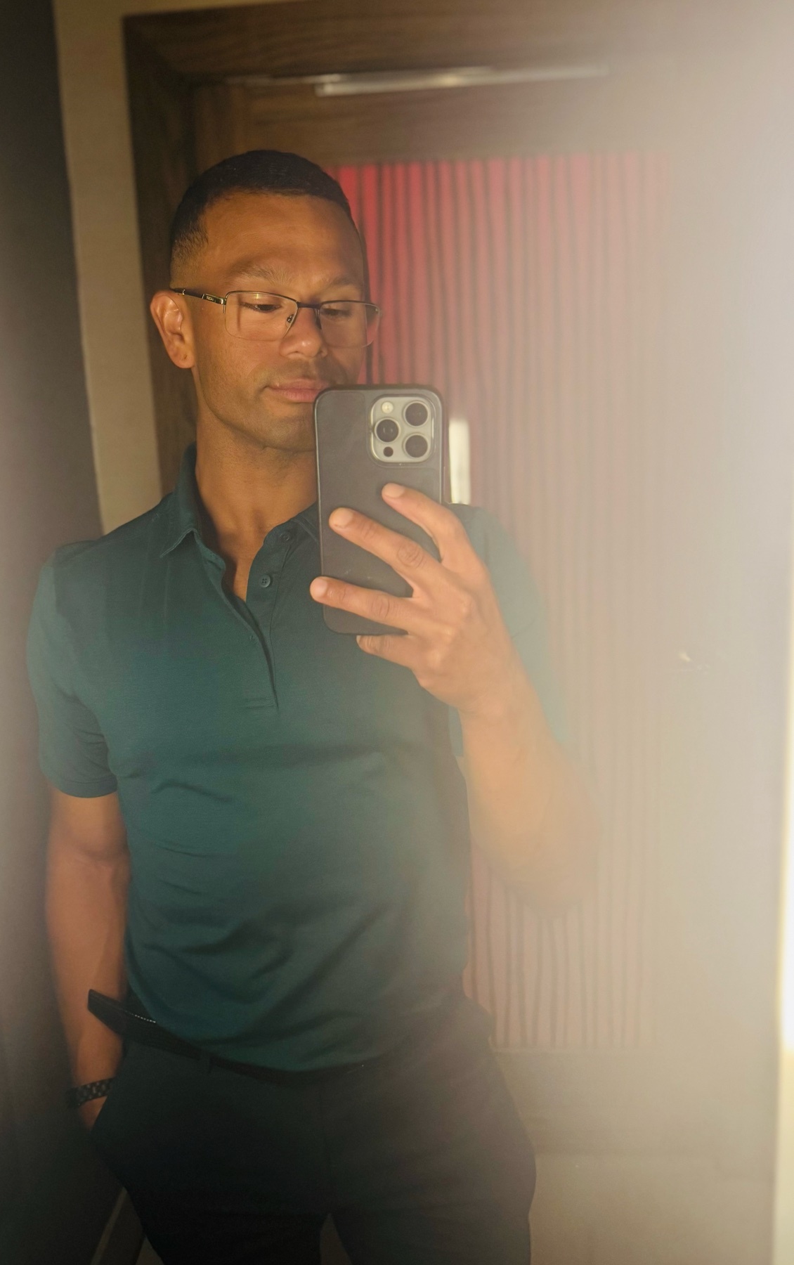 A man takes a mirror selfie, showcasing his outfit. He wears glasses and a polo shirt.