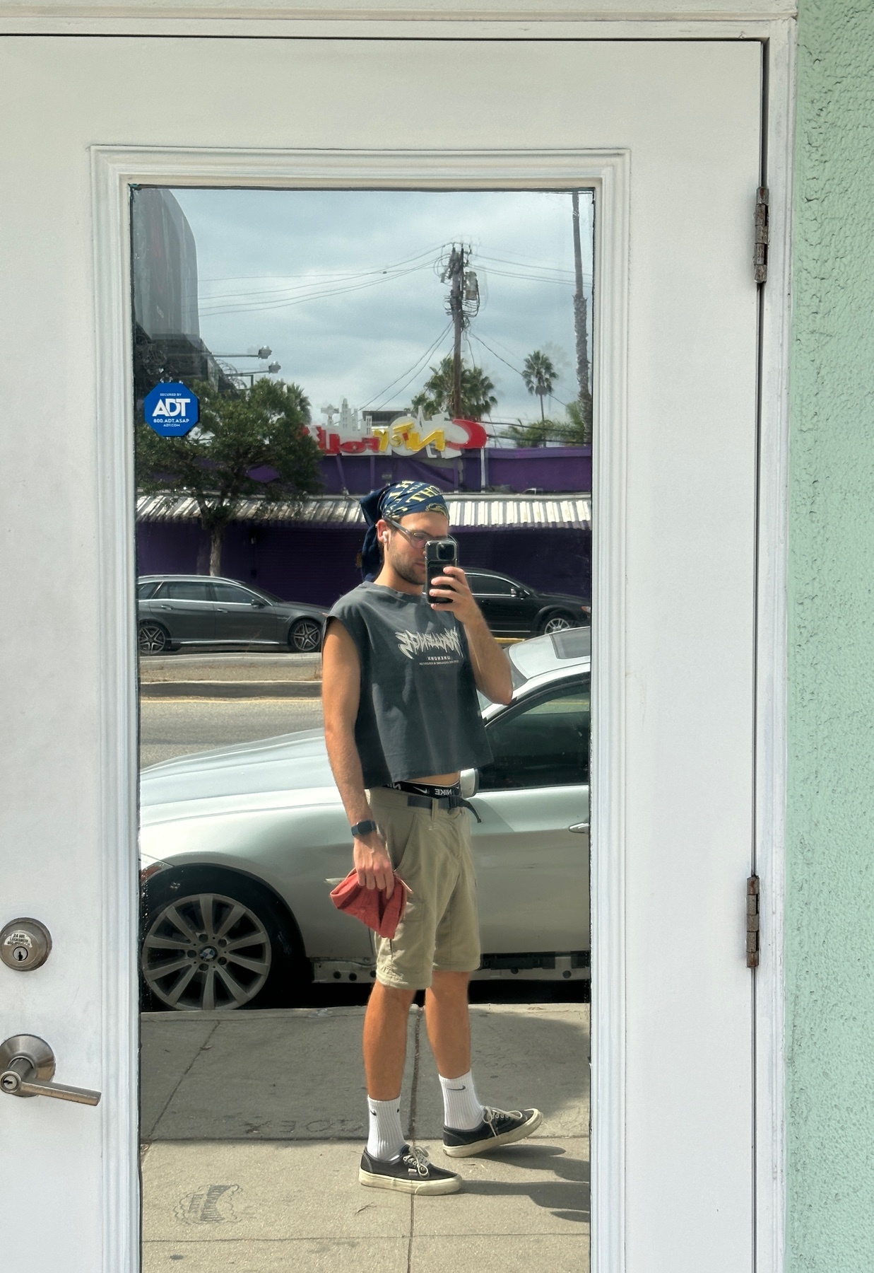 A man takes a mirror selfie in a stylish outfit. He is wearing a crop top, shorts, and a bandana.