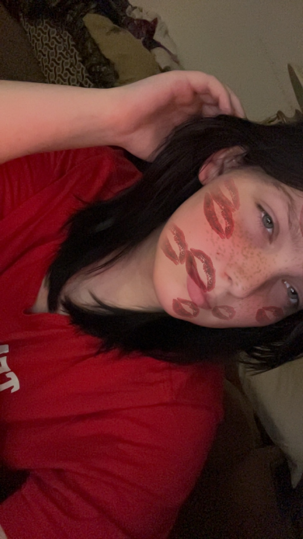 A selfie of a young woman with kiss marks on her face. She is wearing a red shirt.
