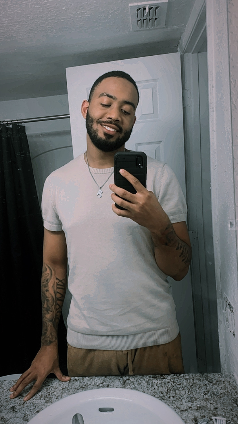 A man takes a mirror selfie in a bathroom. He is smiling and wearing a t-shirt.