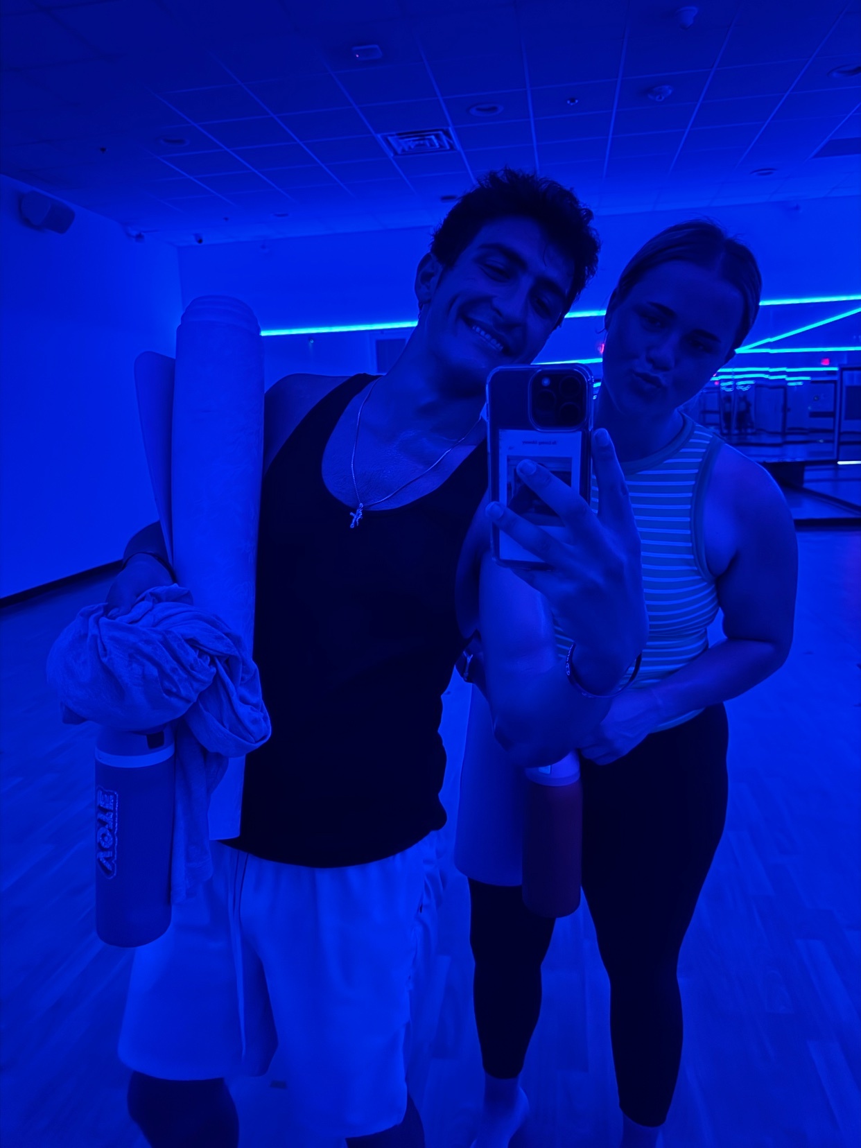 A couple takes a selfie in a dimly lit gym with blue lighting. The man on the left is wearing a black tank top and white shorts, holding a yoga mat and towel. The woman on the right is wearing a striped tank top and dark leggings.