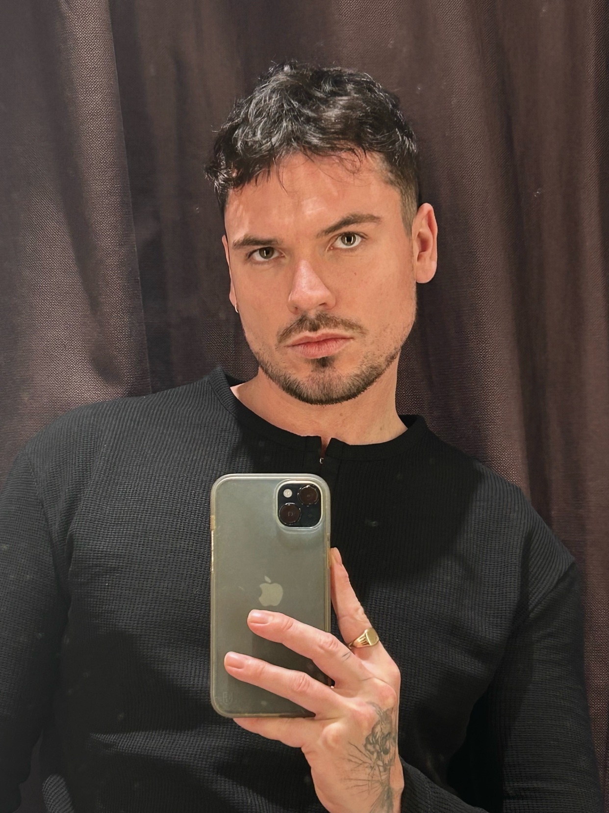 A man with dark hair and stubble takes a selfie in front of a brown curtain. He is wearing a black long-sleeved shirt and a gold ring on his left hand.