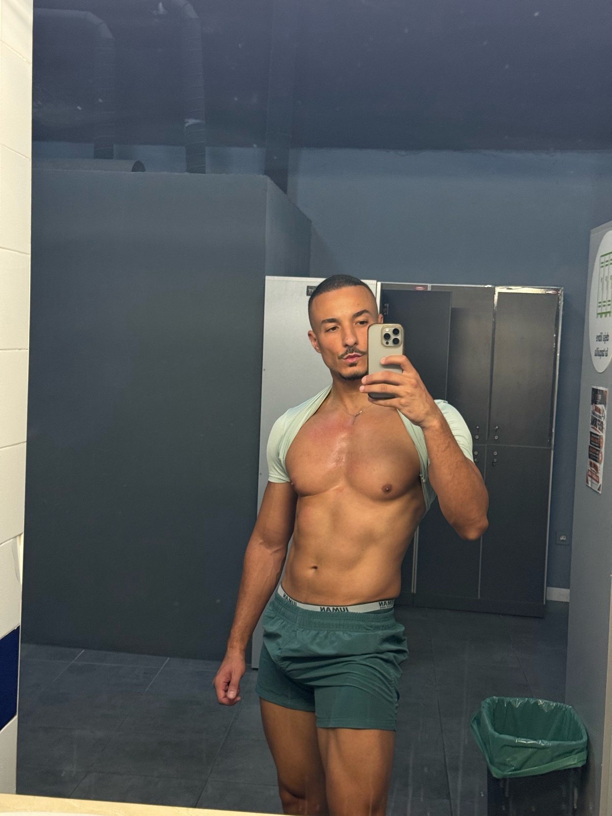 A man takes a mirror selfie in a locker room, wearing short shorts and a t-shirt draped over his shoulders. He has a muscular physique and appears to be in a gym setting.