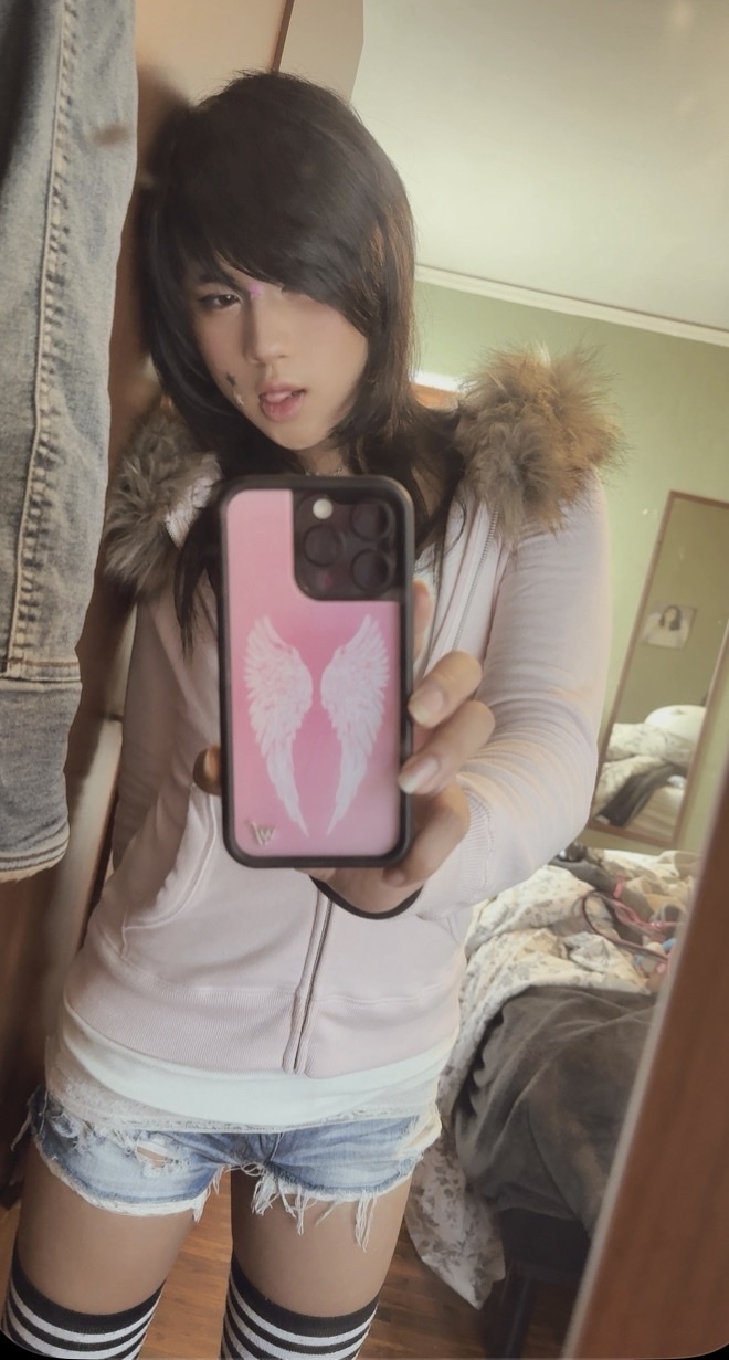 A person takes a mirror selfie, showcasing their outfit. They are wearing a pink jacket, denim shorts, and thigh-high socks.