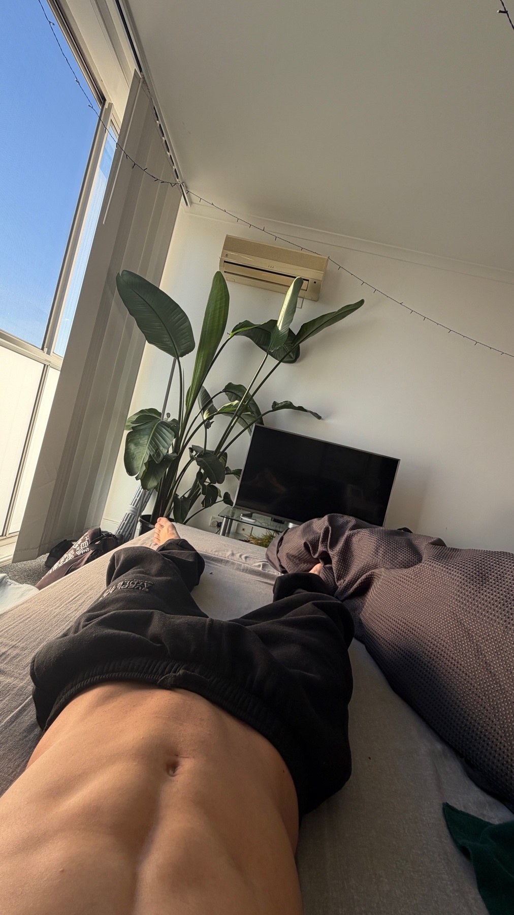 A close-up, low-angle shot of a man's toned abs and legs as he lies on a bed. The room is bright with natural light streaming in from a large window.