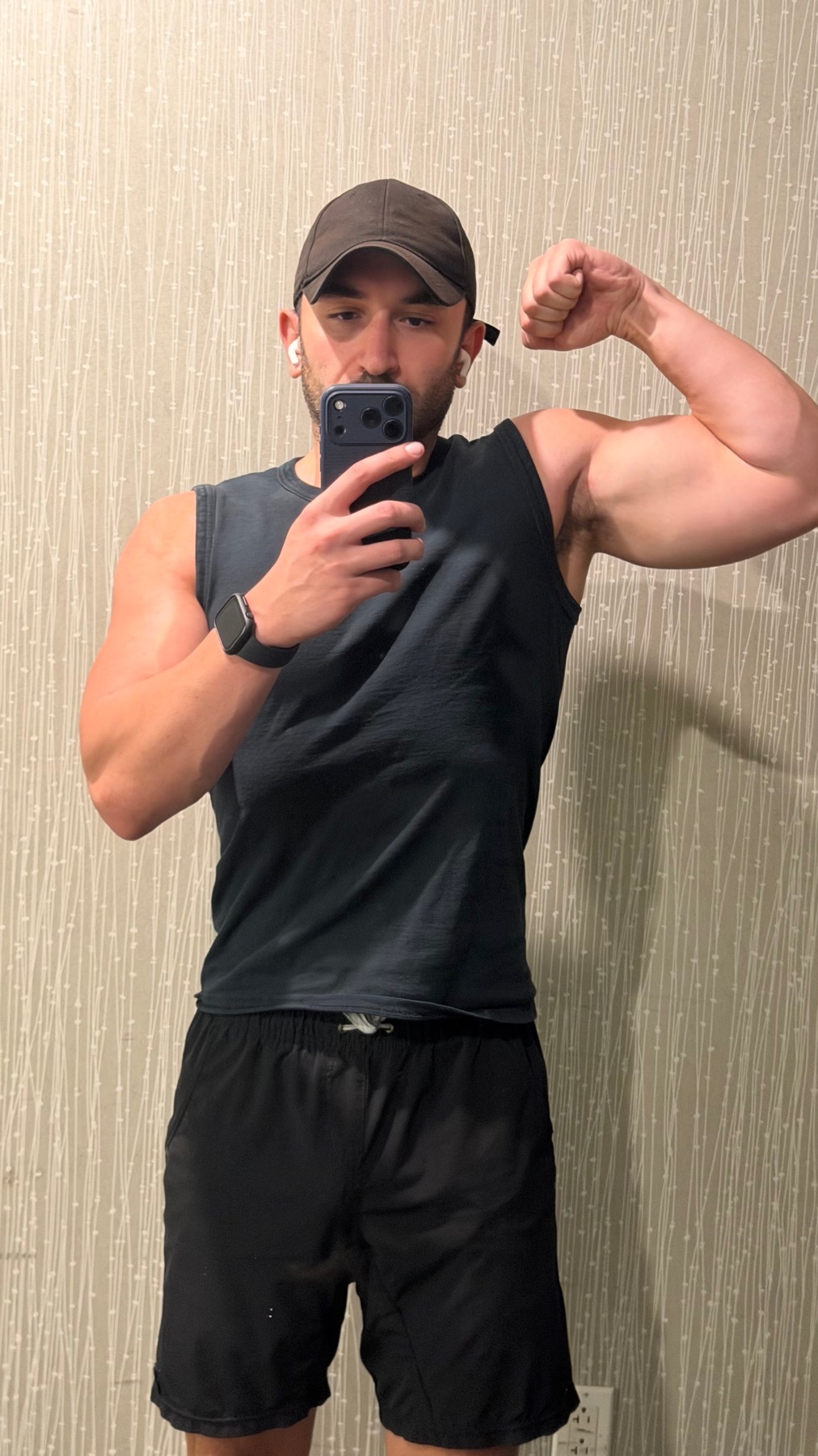 A man in a black tank top and shorts takes a selfie while flexing his bicep. He is wearing a baseball cap and earbuds.