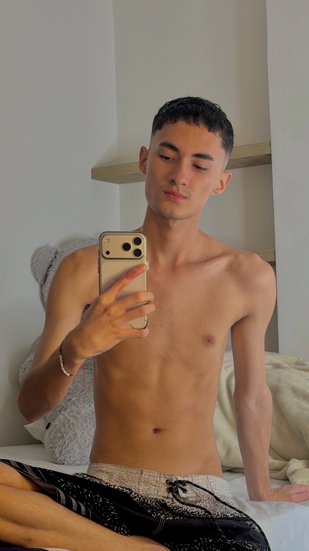 A young man with short, dark hair and tan skin takes a selfie in bed. He is shirtless and wearing patterned shorts.