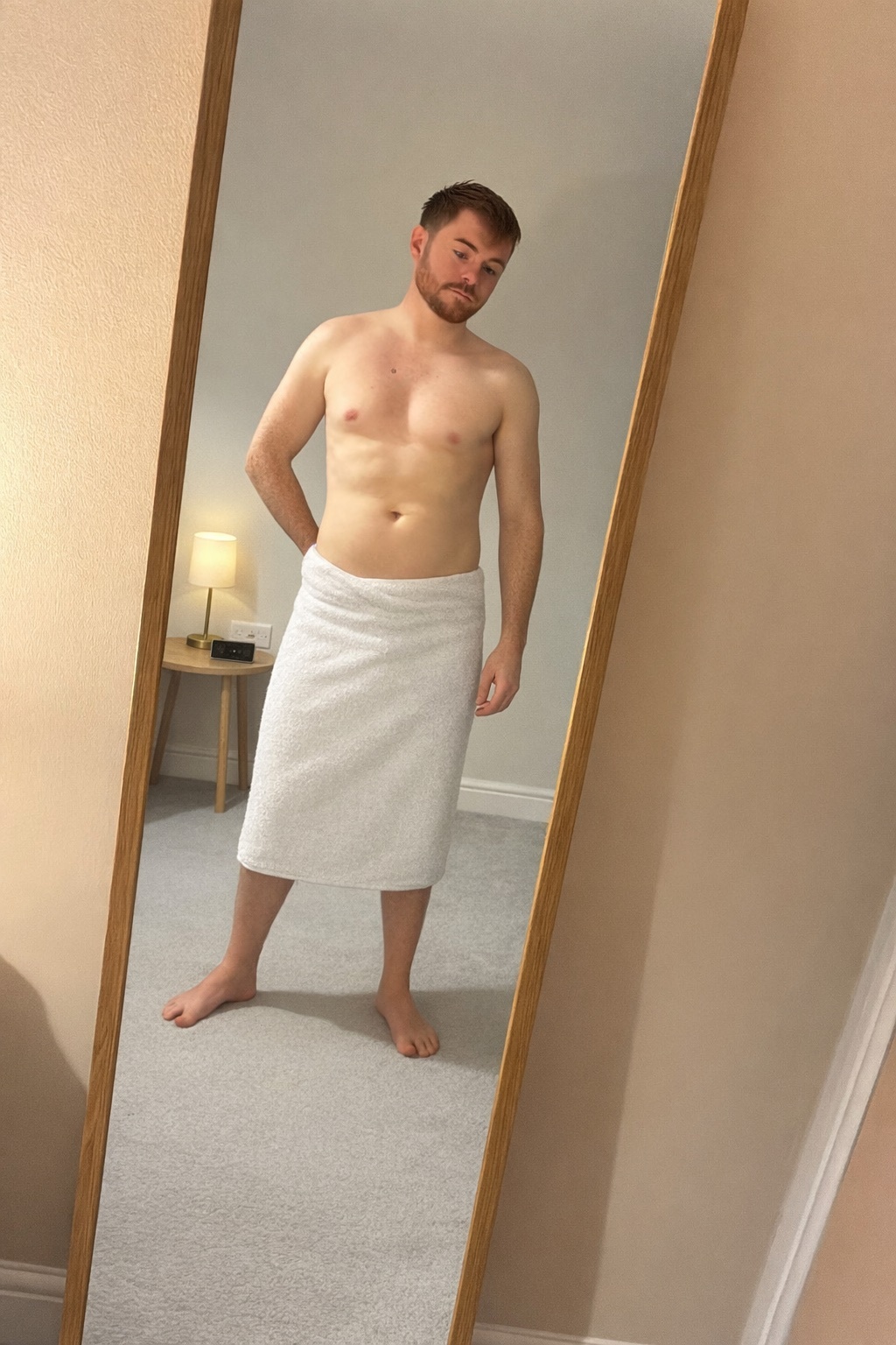 A man stands shirtless in front of a mirror, wrapped in a towel. The photo is taken in a bedroom.