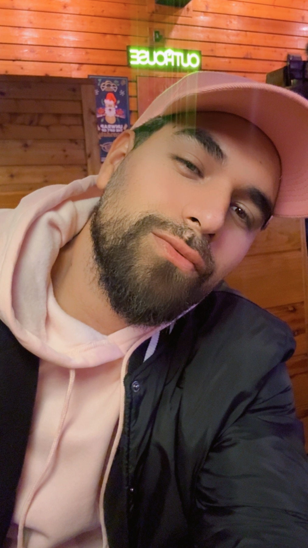 A man poses for a selfie, wearing a pink cap and hoodie. He has a beard and dark hair.