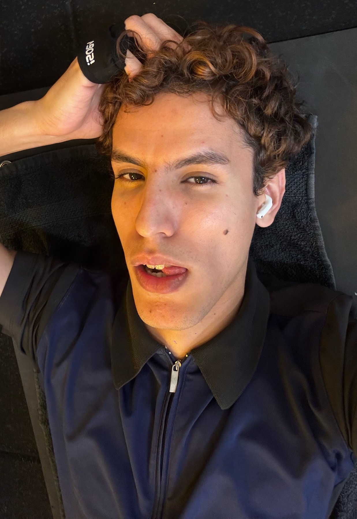 A man with curly brown hair and earbuds in his ears lies on a gym bench, sticking his tongue out.