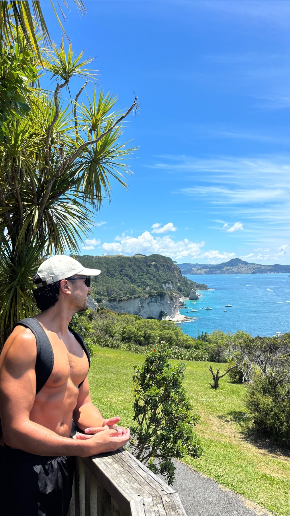 A man stands shirtless, overlooking a scenic coastal landscape. The photo captures a summer day with blue skies and ocean views.