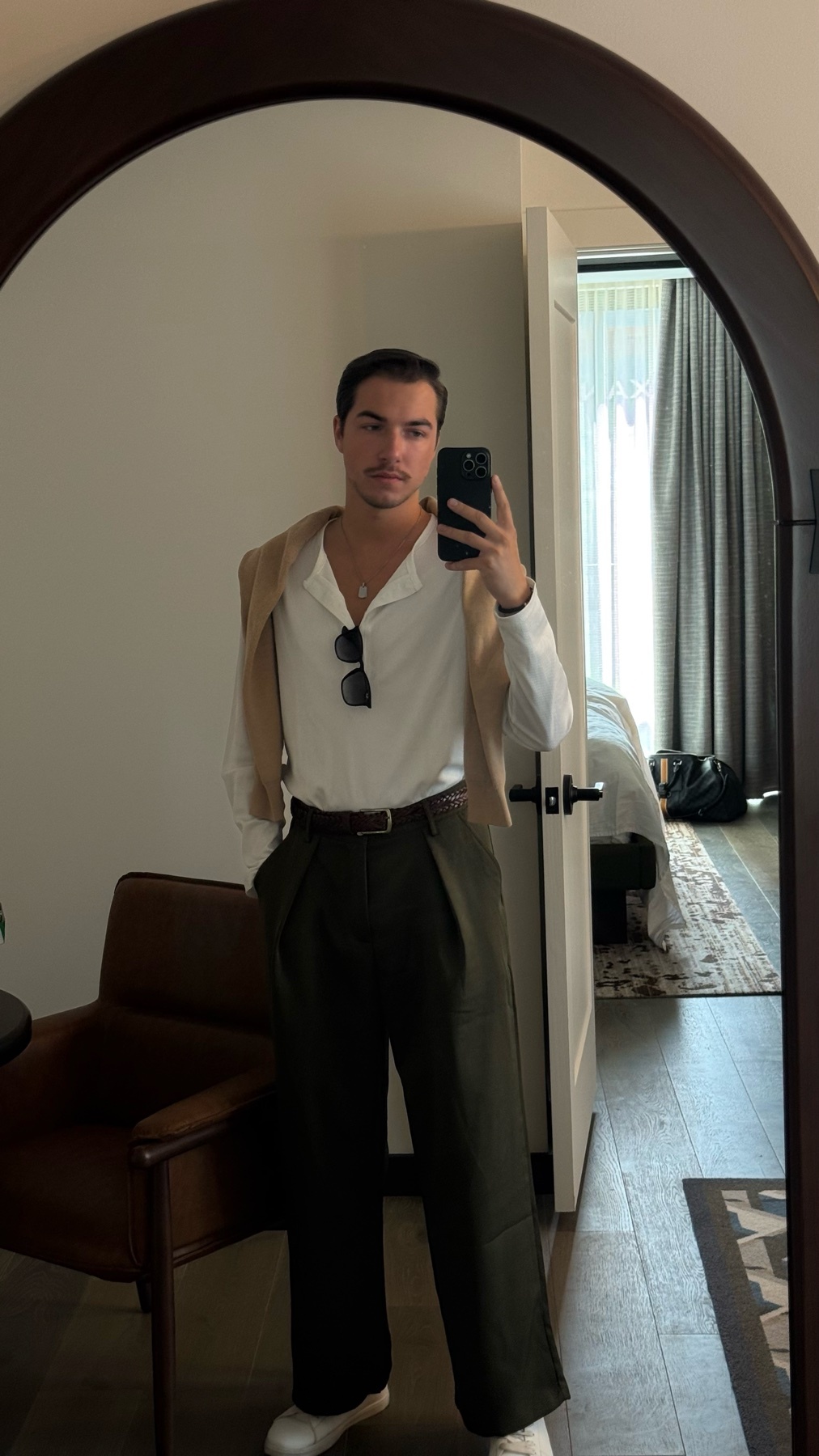 A man takes a mirror selfie in a hotel room, wearing a white button-down shirt, dark green pants, and a tan sweater draped over his shoulders.