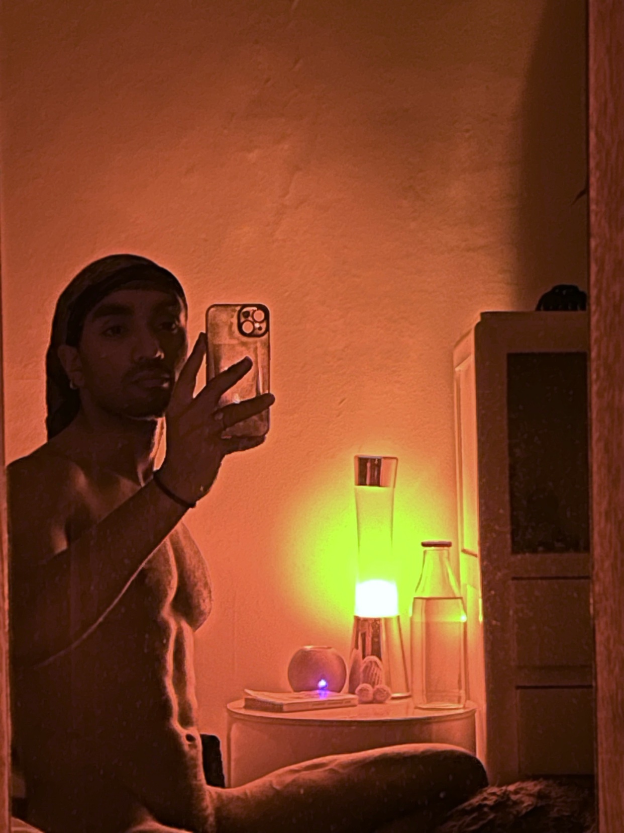 A man takes a shirtless mirror selfie in a dimly lit room. The photo features a lava lamp and other decorative items.