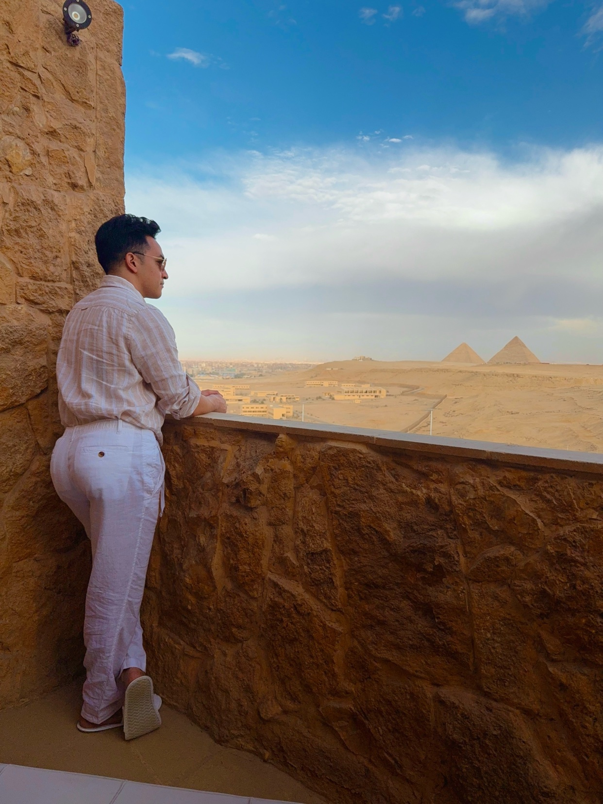 A man stands on a balcony, gazing at the pyramids in the distance. The photo captures a scenic view of the pyramids under a blue sky.