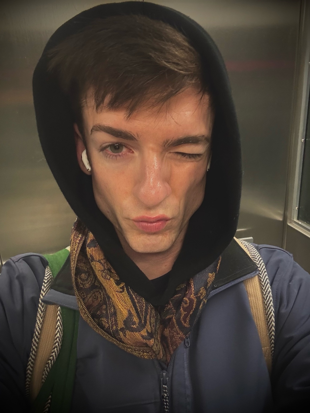 A man in a hoodie winks at the camera. He is wearing a scarf and earbuds.