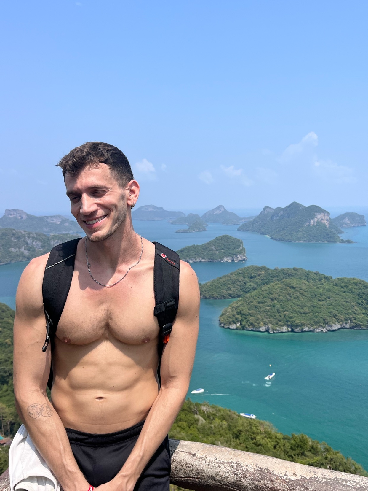 A man poses shirtless with a backpack, overlooking a scenic ocean view in Thailand. The photo captures a moment of travel and fitness.