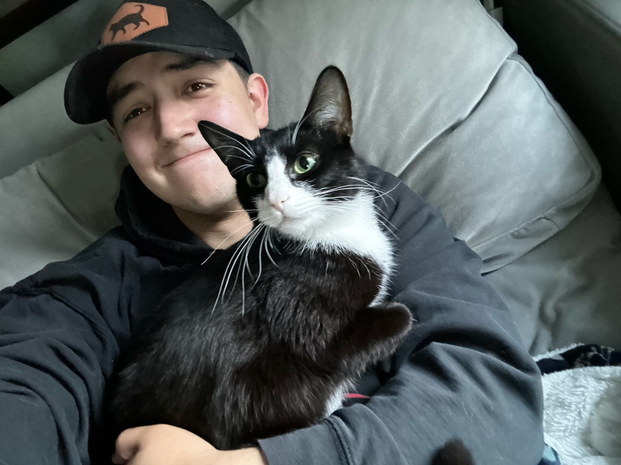 A young man wearing a black baseball cap and hoodie smiles as he holds a black and white tuxedo cat in his arms. The cat has striking green eyes and looks curiously off to the side.