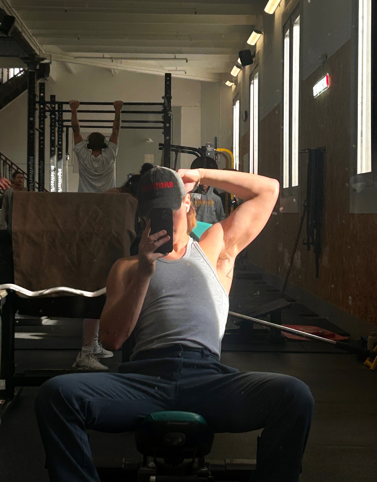 A man takes a mirror selfie at the gym, flexing his arm. He is shirtless and wearing a tank top and a cap.
