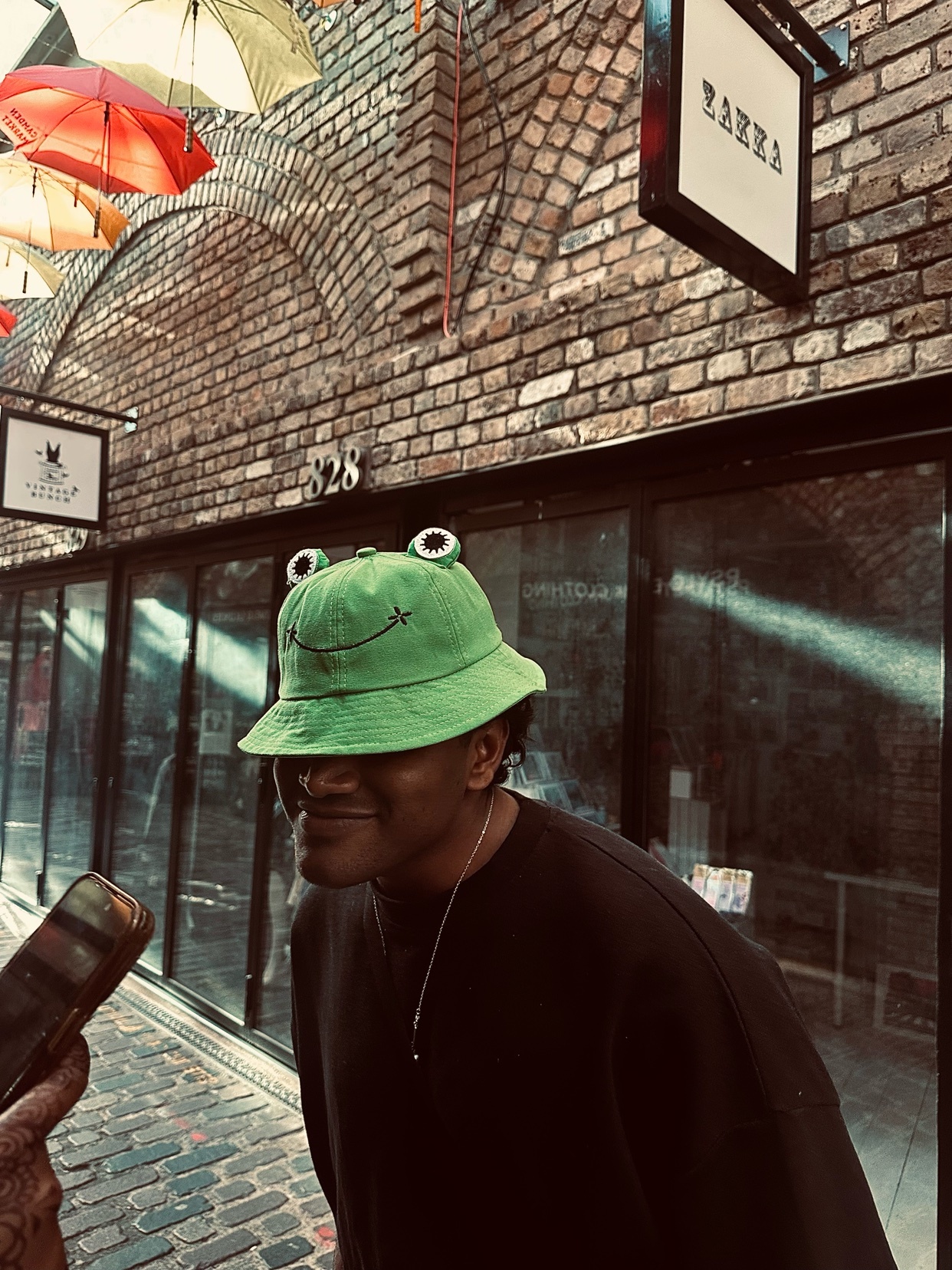 A man wearing a green frog bucket hat smiles in an urban setting. The photo captures a moment of casual style and street fashion.