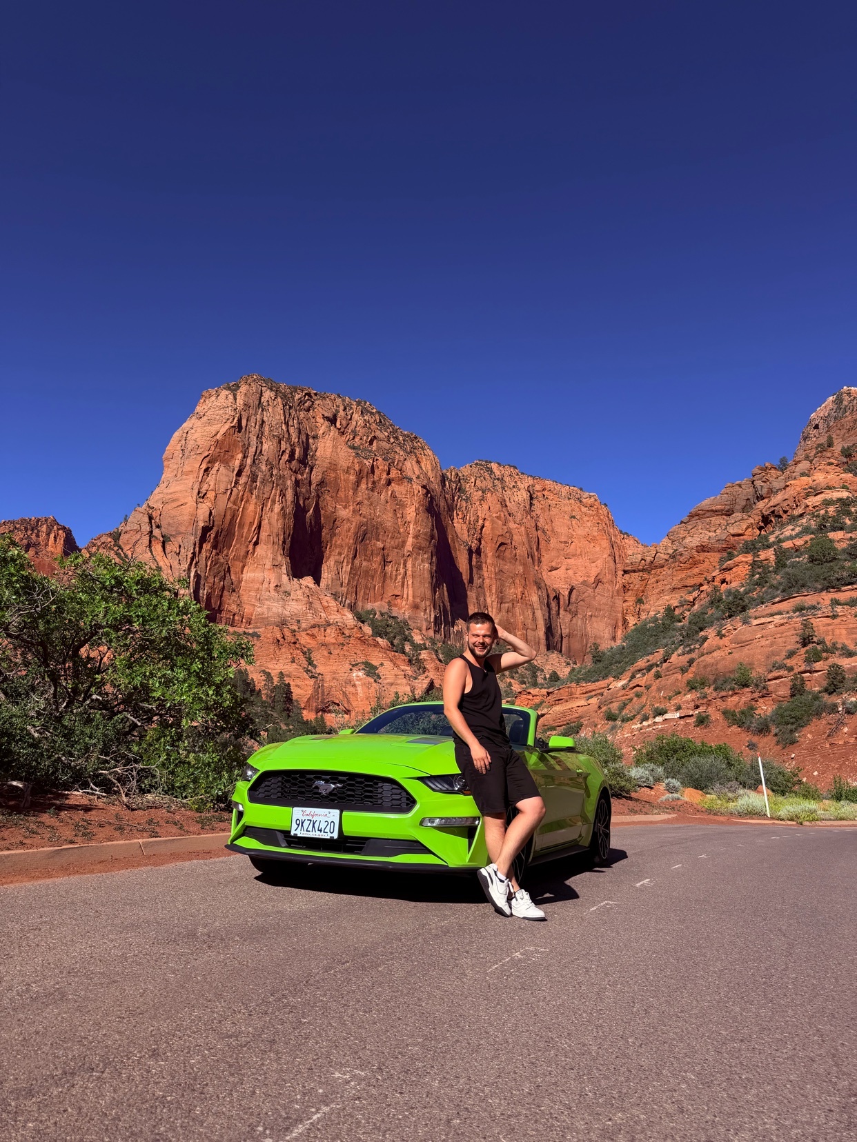 A man poses confidently next to a bright green Ford Mustang with a stunning red rock mountain landscape in the background under a clear blue sky.
