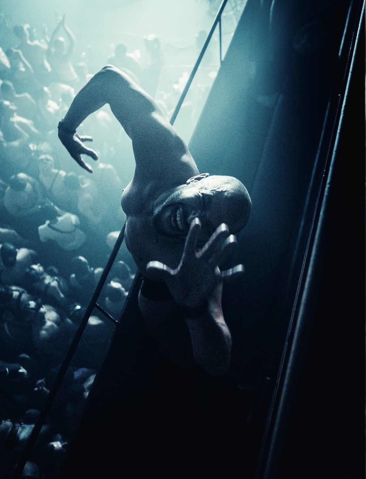 A terrifying, pale creature with sharp teeth and claws climbs upwards, reaching out with its hands towards a blurred crowd of people below. The scene is dark and dramatic, illuminated by a single spotlight, creating a sense of intense horror and susp