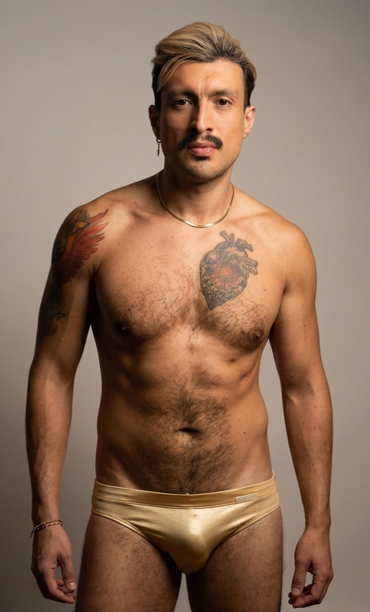A man with a mustache and tattoos poses shirtless in gold bikini briefs. He has chest hair and is wearing a necklace.